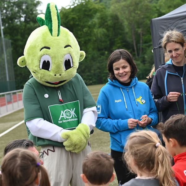 Person im grünen Jolinchen-Kostüm mit AOK-Logo neben Kristine Weinhold von der Stiftung Anstoß zum Leben und Nadine Dietrich vom Kreissportbund, die Kindern Medaillen überreichen auf Sportplatz